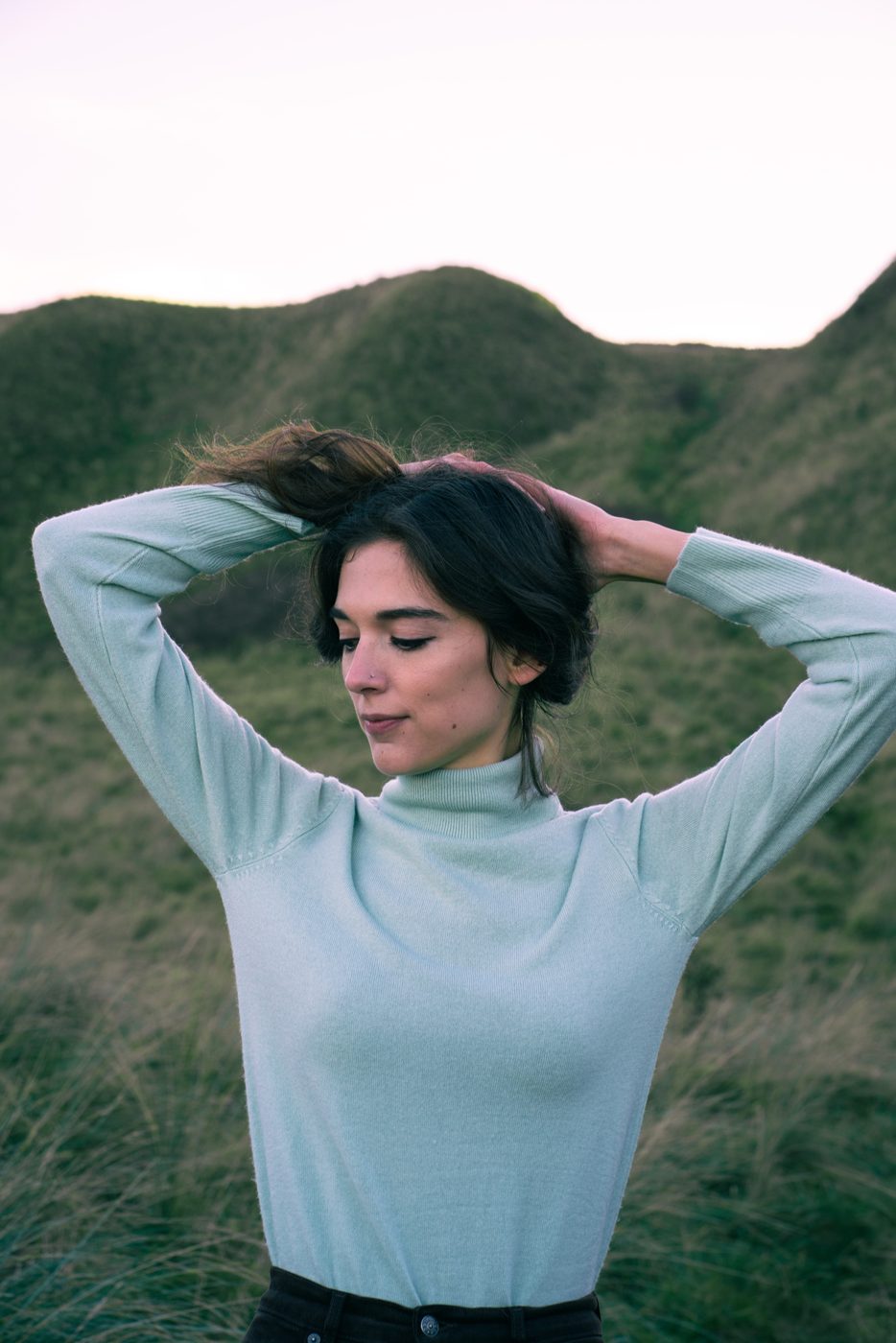 Hands in hair in a pale blue turtleneck on a hillside